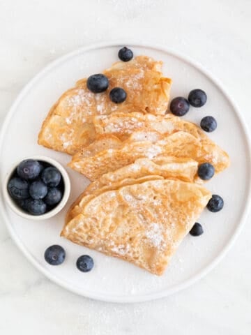 Overview of a plate with several folded vegan crepes topped with fresh blueberries and powdered sugar.