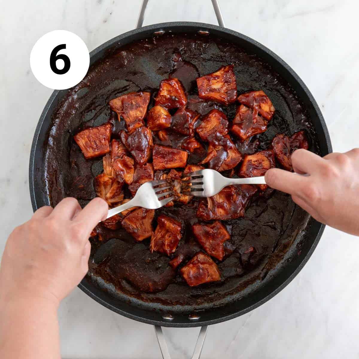 Sauteéd jackfruit in a skillet with BBQ sauce, while two hands shred it with forks.