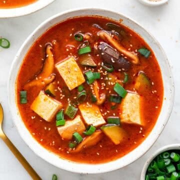 Two bowls of spicy Korean tofu soup, surrounded by bowls of toppings and a spoon.