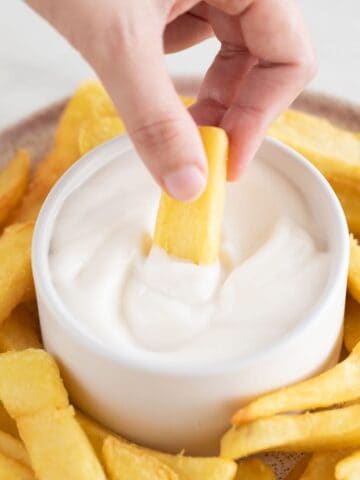Photo of a dish with French fries and a small bowl with vegan aioli and a hand dipping a fry into the sauce.