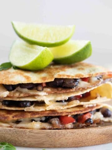 Side photo of a pile of vegan quesadillas onto a wooden board with some lime wedges